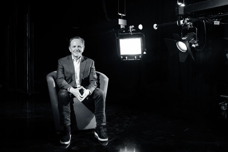 Black and white photo of Brian Walsh sitting on a chair smiling at the camera