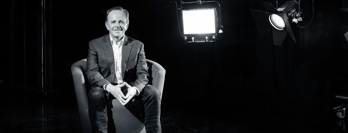 Black and white photo of Brian Walsh sitting on a chair smiling at the camera