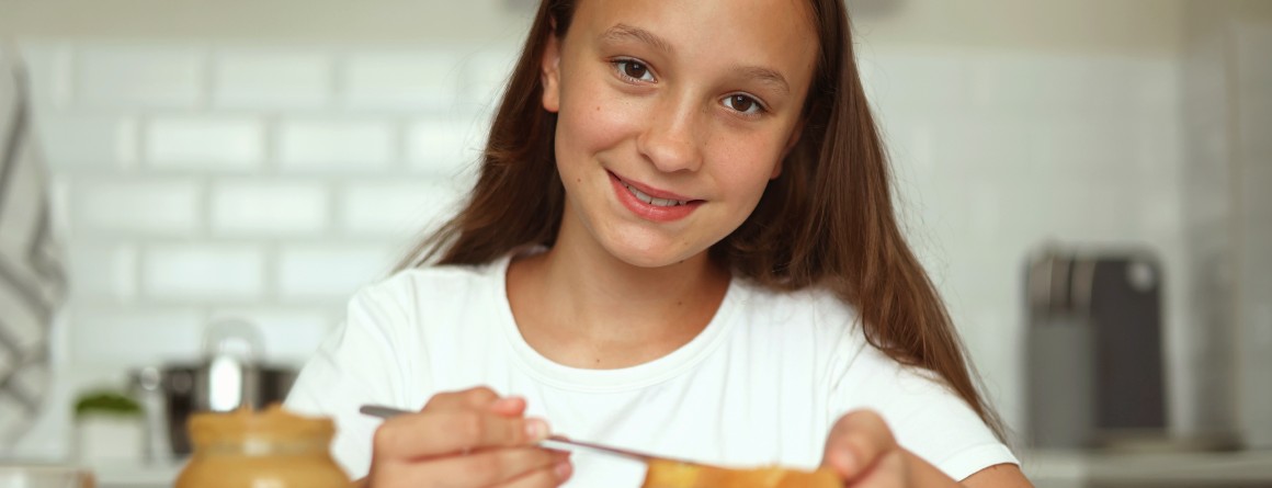 A young woman with brown hair spreads peanut butter over a piece of toast. She wears a white T-shirt and smiles to the camera.