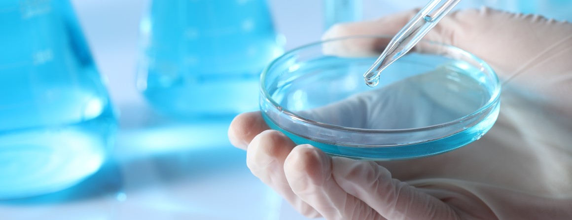 A petri dish sits in a researcher's hands, which wear white latex gloves. A pipette tool sits in the dish, dripping a small amount of liquid. In the background are beakers of light blue liquid.