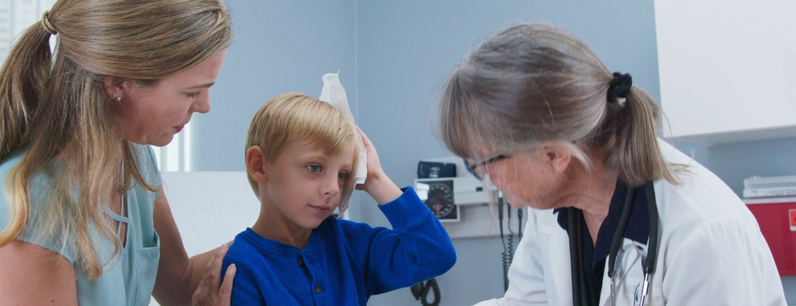 A concussed child with an ice pack on their head, being attended to by a doctor and their mother