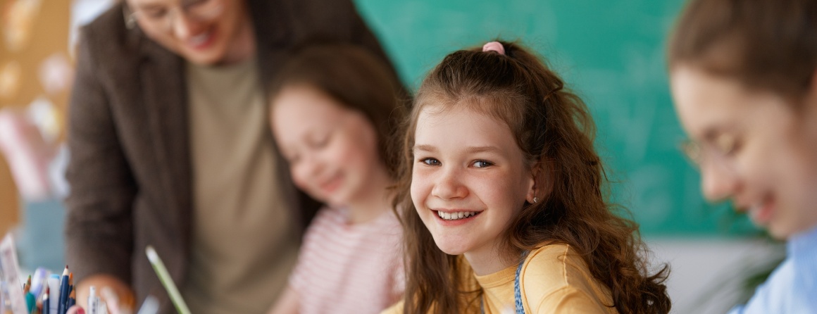 An adolescent girl at school, smiling at the camera. She holds a pencil in her right hand and wears her auburn hair in a half-up half-down style. There are other students and a teacher on either side of her.