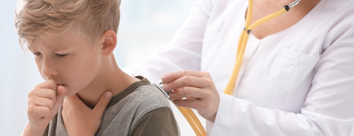 A fair haired boy of primary school age coughs while holding a fist to his mouth. A doctor assesses his lungs using a stethoscope.