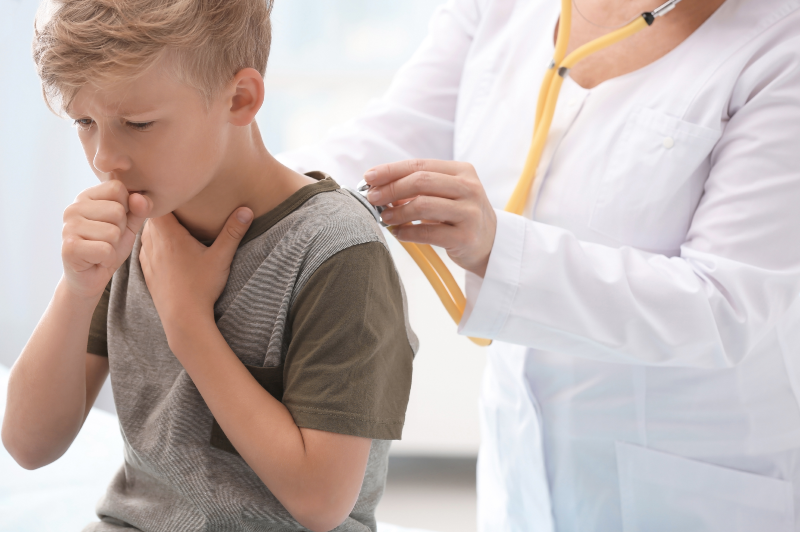 A fair haired boy of primary school age coughs while holding a fist to his mouth. A doctor assesses his lungs using a stethoscope.