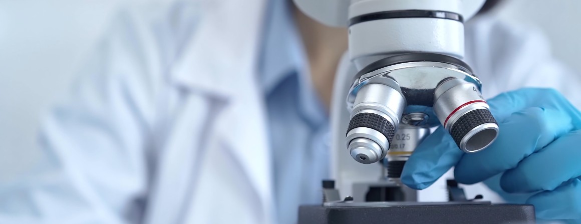 A researcher examines an object under a microscope. Their face is out of shot, but we can see their hand adjusting a lens. They wear a white lab coat and blue nitrile gloves.