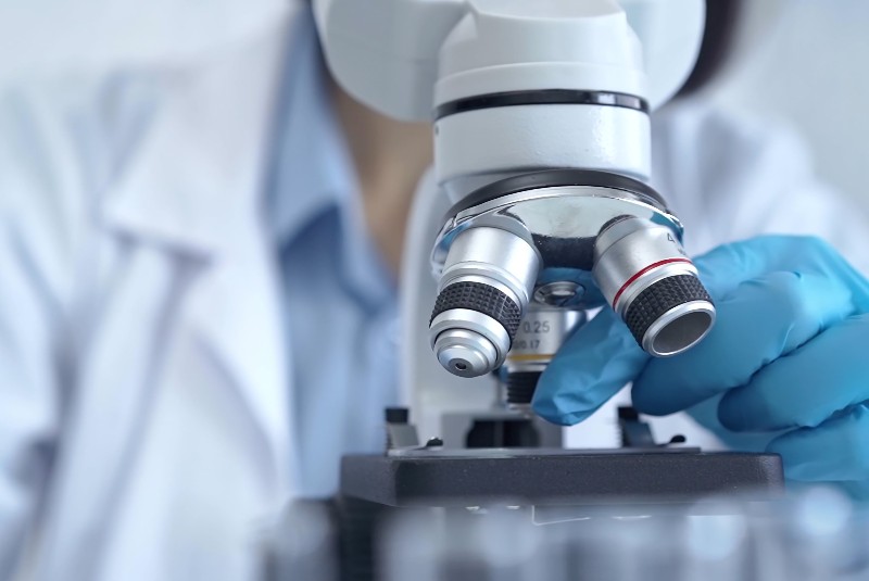 A researcher examines an object under a microscope. Their face is out of shot, but we can see their hand adjusting a lens. They wear a white lab coat and blue nitrile gloves.
