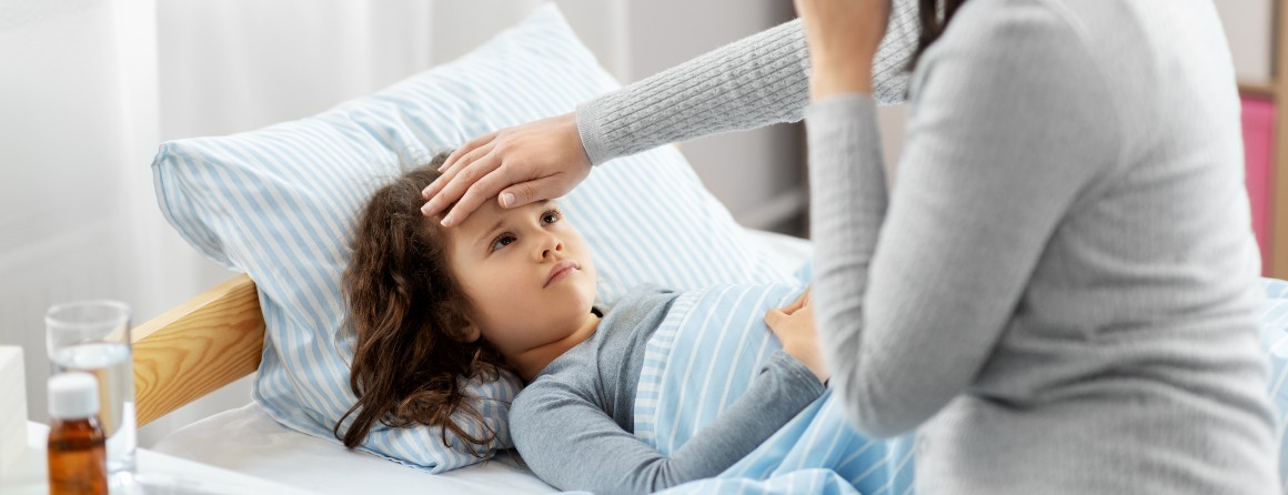 A primary school aged girl is sick in bed, her mother places a hand on her daughter's forehead. She wears a grey sweater and has dark, shoulder-length hair.