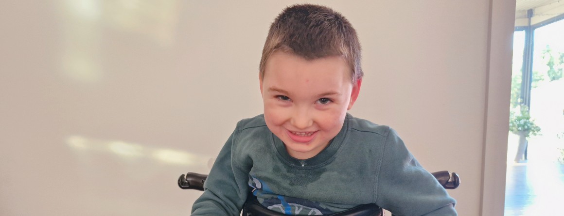 A young boy with light brown hair smiles to the camera. He balances upright using his walker and wears a light blue jumper.