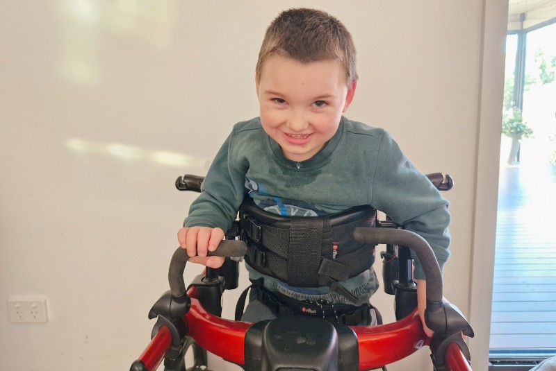 A young boy with light brown hair smiles to the camera. He balances upright using his walker and wears a light blue jumper.