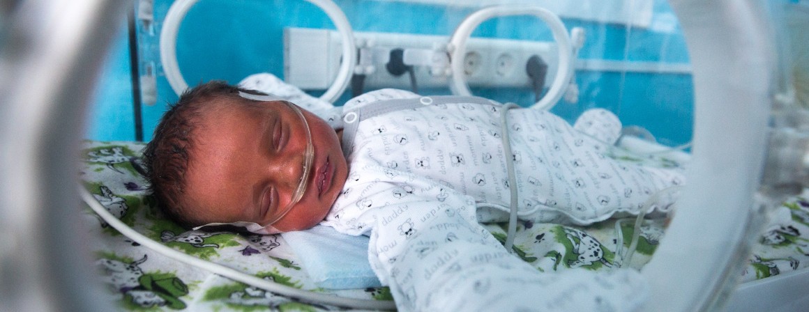 A newborn baby, connected to breathing support, naps inside a clear incubator, which has small windows on the sides.