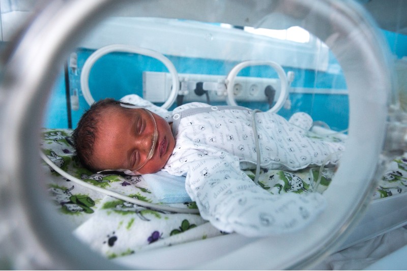 A newborn baby, connected to breathing support, naps inside a clear incubator, which has small windows on the sides.