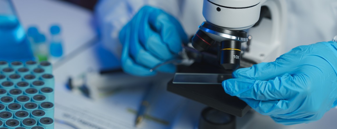 A researcher examines an object under a microscope. They wear blue, nitrile gloves and a white lab coat. Their face is not visible.