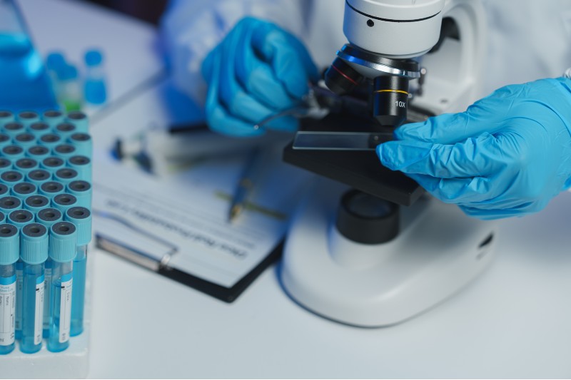 A researcher examines an object under a microscope. They wear blue, nitrile gloves and a white lab coat. Their face is not visible.