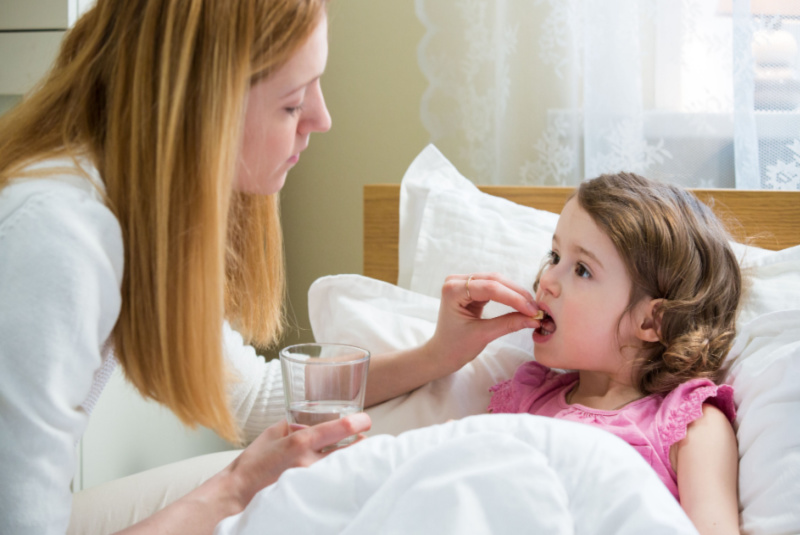 Worried mother giving glass of water to her ill kid. Sick child with high fever laying in bed and taking a medicine. Hand on forehead