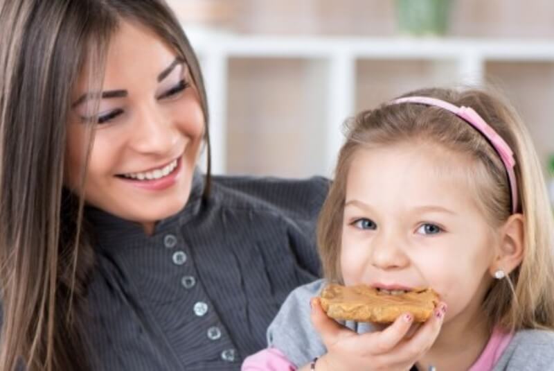 Smiling woman watching young girl eat a peanut butter sandwich
