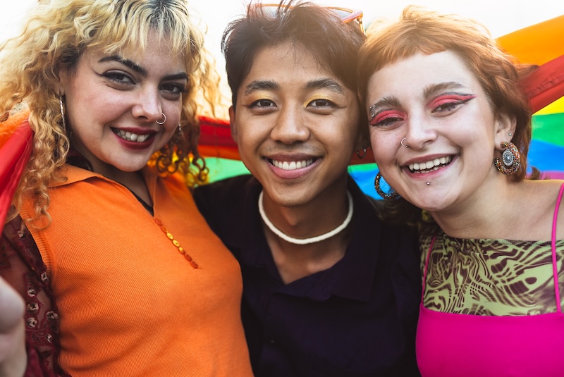 3 LGBTQIA+ persons arm in arm holding a pride flag